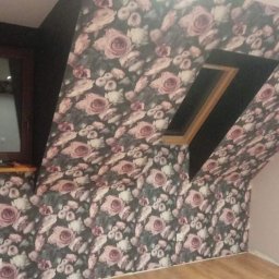 Rad-rud-system - Pokój na poddaszu po tapetowaniu, ściany i sufit pokryte tapetą w ciemne róże, widoczne okno dachowe i okno w ścianie szczytowej, podłoga drewniana.