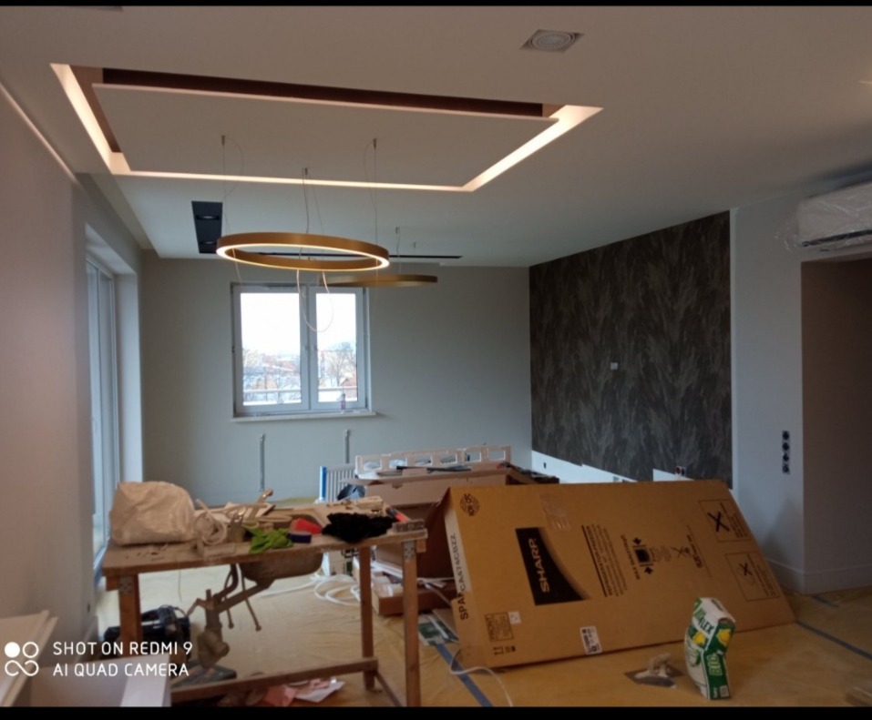 Wnętrze w trakcie prac wykończeniowych: sufit podwieszany z oświetleniem LED, okrągłe lampy wiszące, ściana z tapetą o wzorze marmuru, narzędzia i materiały budowlane na stole roboczym, karton po...