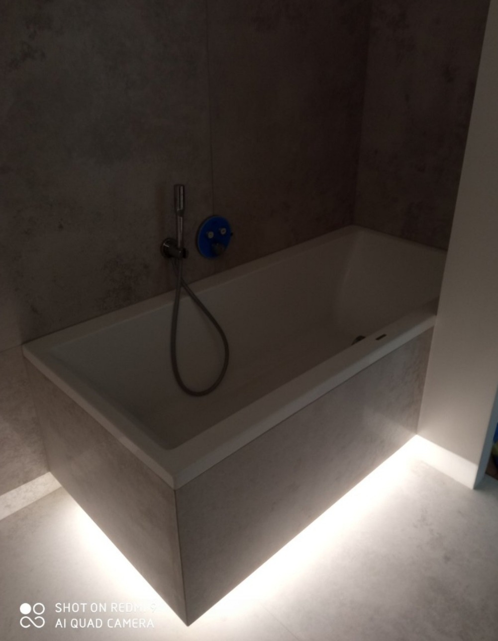 Nowoczesna wanna obudowana płytami imitującymi beton, podświetlona listwą LED, z baterią natryskową na ścianie z efektem betonu.