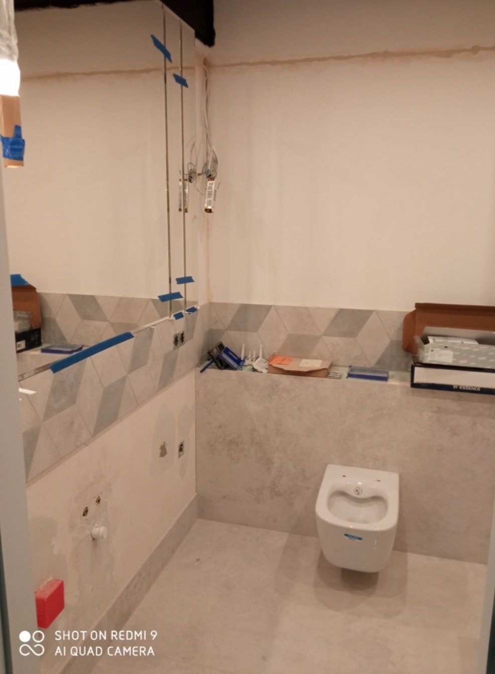 Łazienka w trakcie montażu podwieszanej toalety, widoczne rury, płytki z geometrycznym wzorem na ścianie, oraz narzędzia i materiały budowlane na półce.