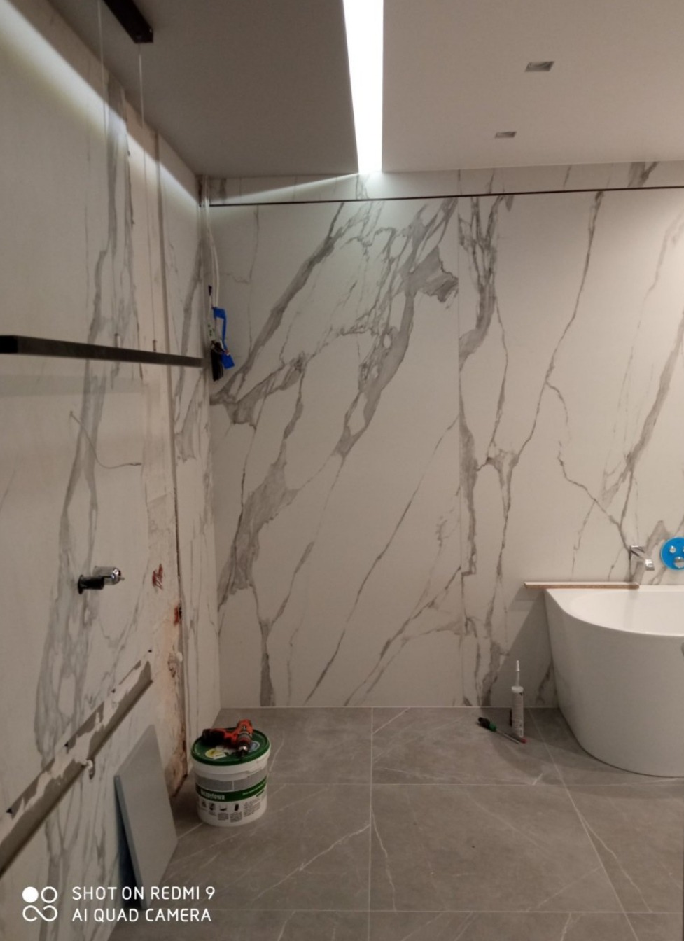 Łazienka w trakcie remontu: płytki imitujące marmur na ścianach, biała wanna wolnostojąca, widoczny wiadro z zaprawą i narzędzia glazurnicze.