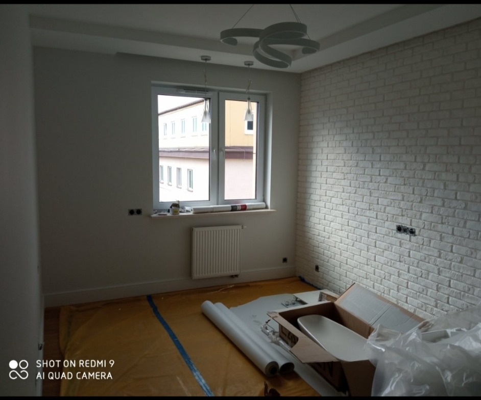 Pokój w trakcie prac wykończeniowych: ściana z białej cegły dekoracyjnej, zabezpieczona podłoga folią, widoczne rolki tapet i karton z elementami wyposażenia.