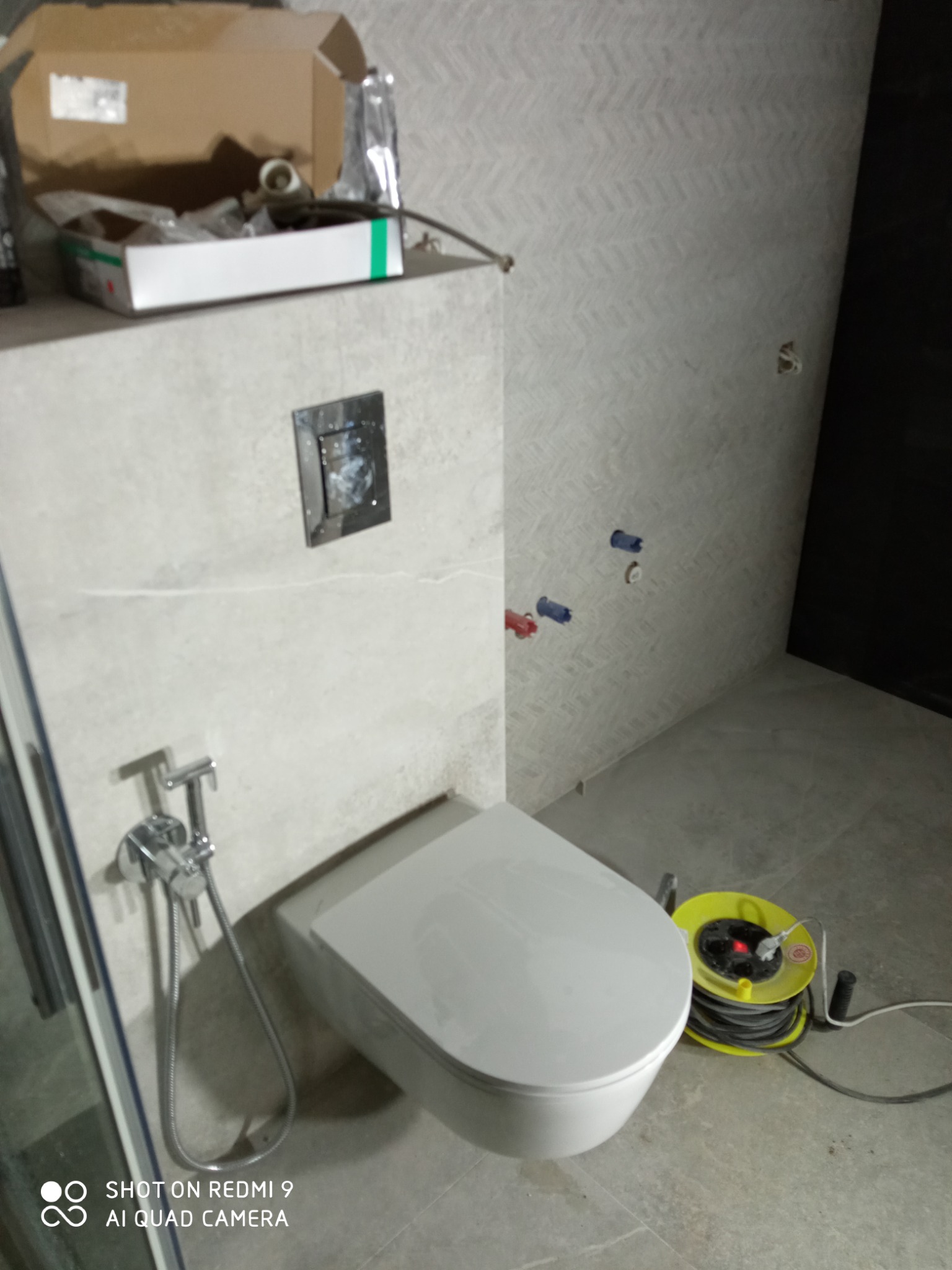 Montaż wiszącej toalety z bidetką w łazience z szarą, wzorzystą tapetą, widoczne przyłącza wodne i zwinięty przedłużacz elektryczny.