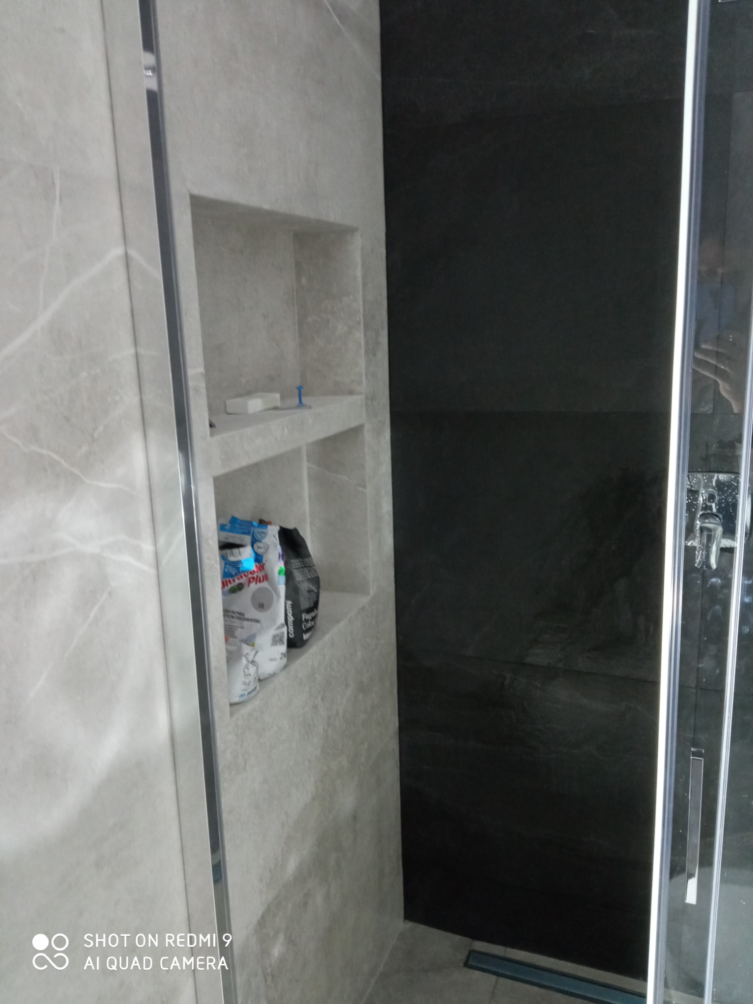 Wykończona łazienka z szarą, marmurkową płytką, wnęka z półkami, na których stoją worki z zaprawą klejową, czarna ściana w kabinie prysznicowej.
