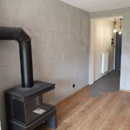 Wnętrze pokoju z wykończonymi szarymi ścianami imitującymi beton, drewnianą podłogą, czarnym piecem wolnostojącym z szybą i fragmentem wejścia do innego pomieszczenia z szarą podłogą w kształcie...