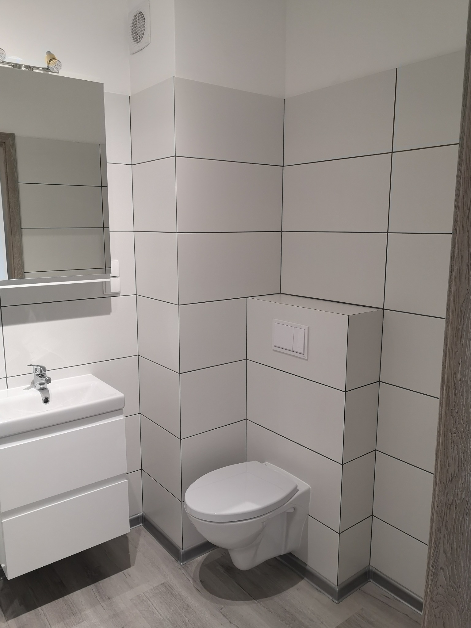 Nowoczesna, minimalistyczna łazienka z białymi, kwadratowymi płytkami na ścianach, podwieszaną toaletą i białą szafką z umywalką na szarych panelach podłogowych.