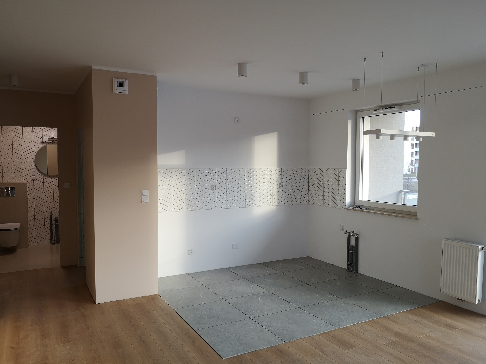 Wnętrze w trakcie prac wykończeniowych: ułożone szare płytki podłogowe w części pomieszczenia, białe ściany z fragmentem dekoracyjnej mozaiki w jodełkę, grzejnik, instalacja wodna przy oknie, widok...