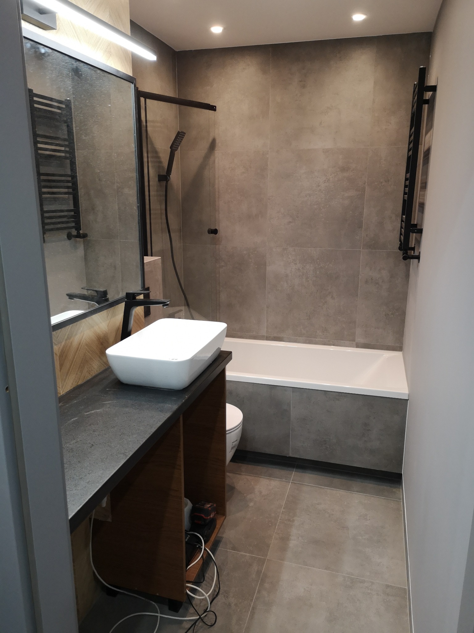 Nowoczesna łazienka z umywalką nablatową, czarną armaturą i szarymi płytkami, widok na wannę i grzejnik drabinkowy.