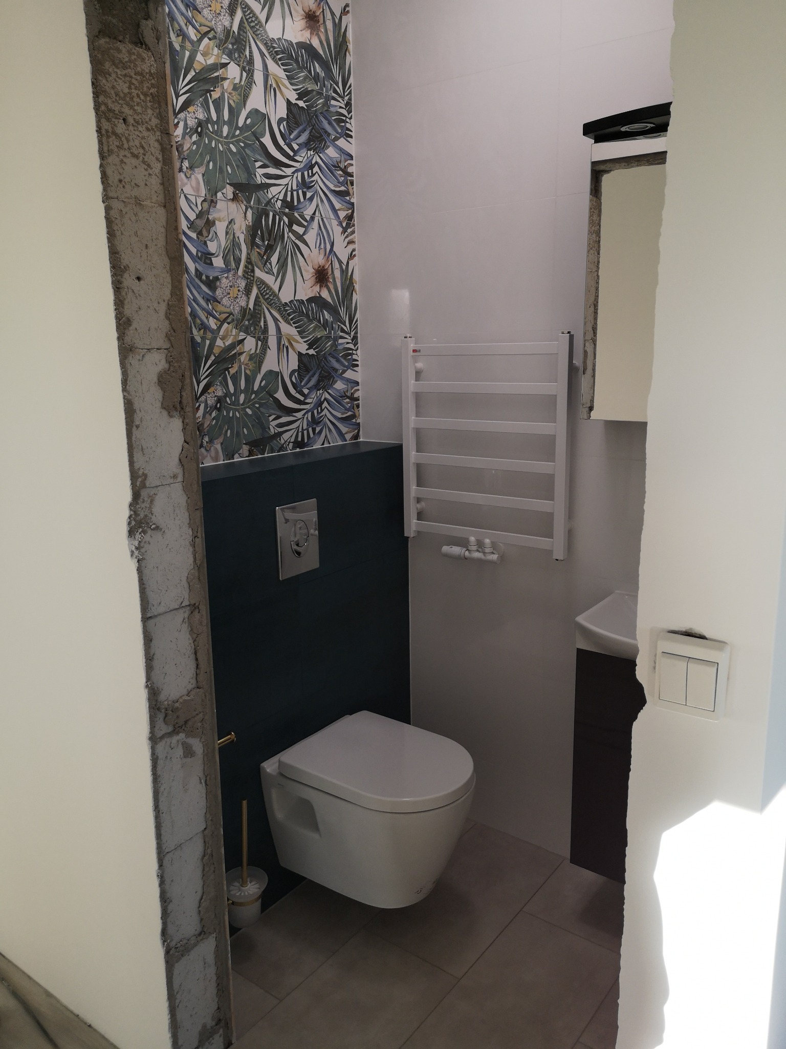Nowoczesna toaleta wisząca w łazience z fragmentem ściany pokrytej tapetą w motywy roślinne, obok grzejnik drabinkowy i umywalka z szafką w ciemnym kolorze, widoczne niedokończone wykończenie...