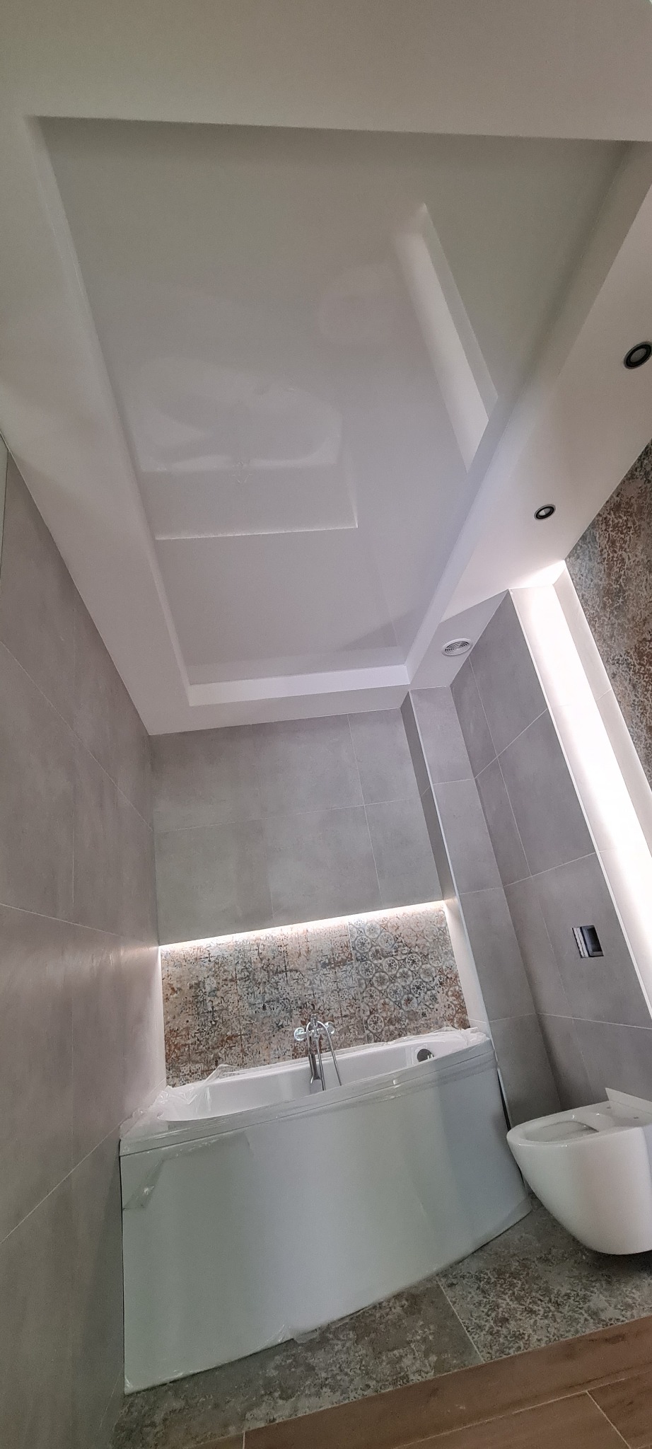 Biały, podwieszany sufit z wbudowanym oświetleniem punktowym w nowoczesnej łazience z szarymi płytkami i podświetlaną ścianą nad wanną, częściowo zabezpieczoną folią ochronną.