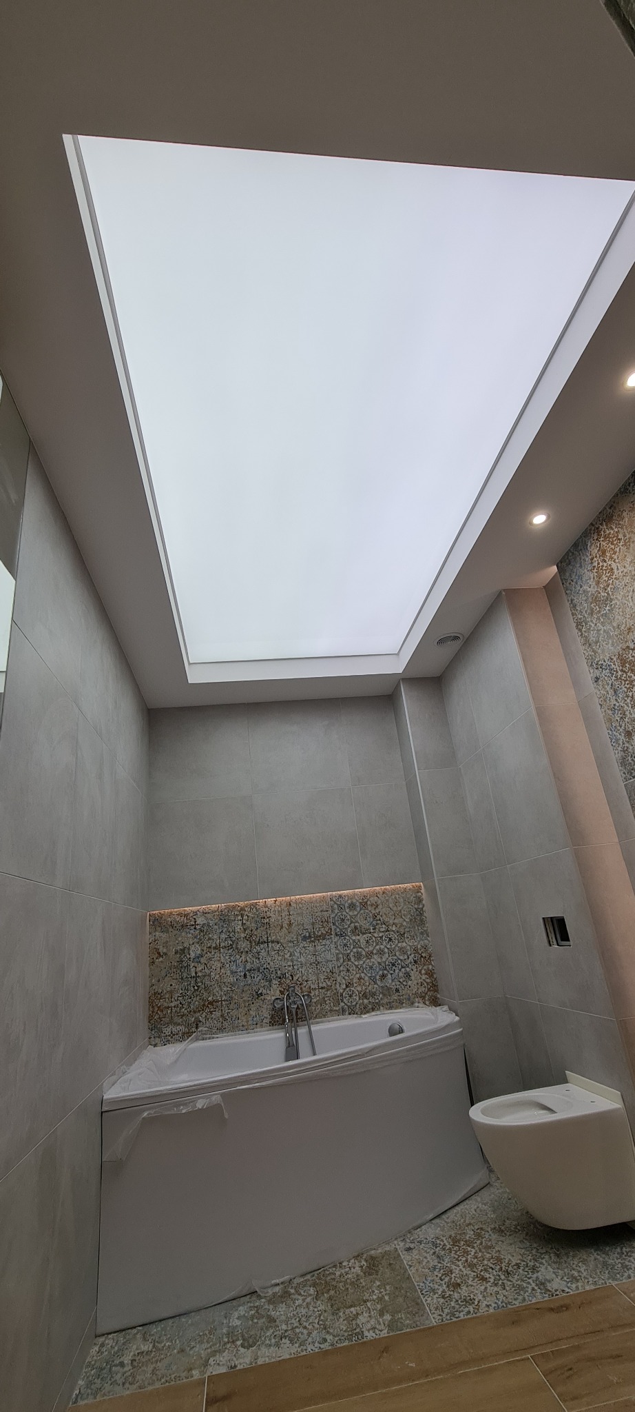 Nowoczesna łazienka z podświetlanym sufitem napinanym, szara glazura ścienna i mozaika nad wanną narożną, widoczna toaleta podwieszana.