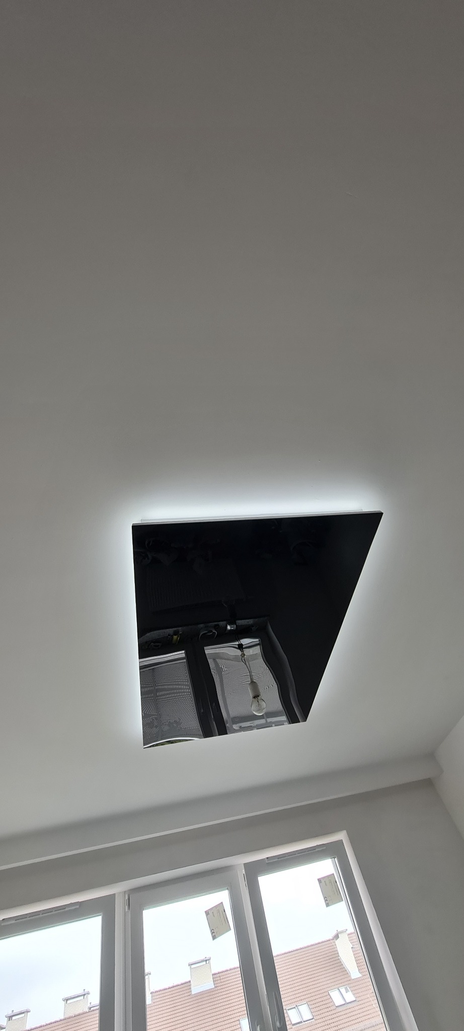 Czarny, kwadratowy sufit napinany z podświetleniem LED, odbijający okno z roletami i żarówkę