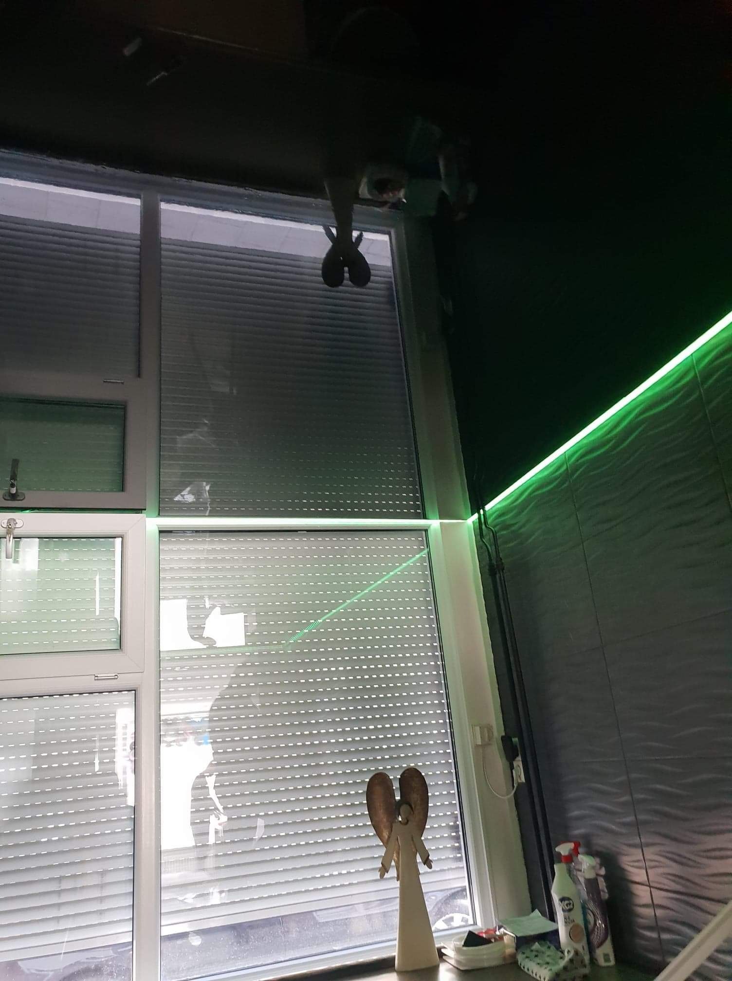 Okna z roletami wewnętrznymi, dekoracyjna figurka anioła na parapecie, ściana z szarej, falistej płytki i zielone oświetlenie LED.