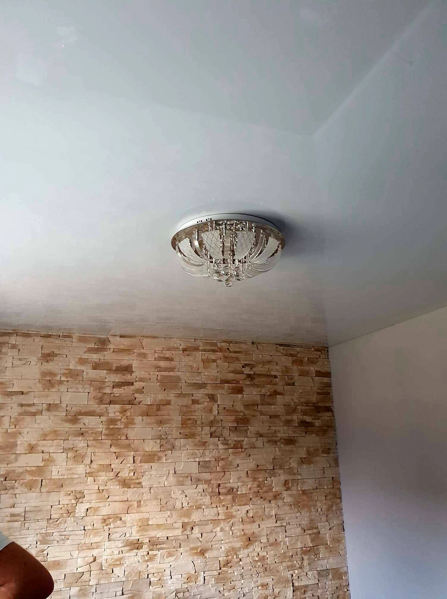 Błyszczący, biały sufit napinany z zamontowanym ozdobnym żyrandolem, ściana wykończona imitacją kamienia.