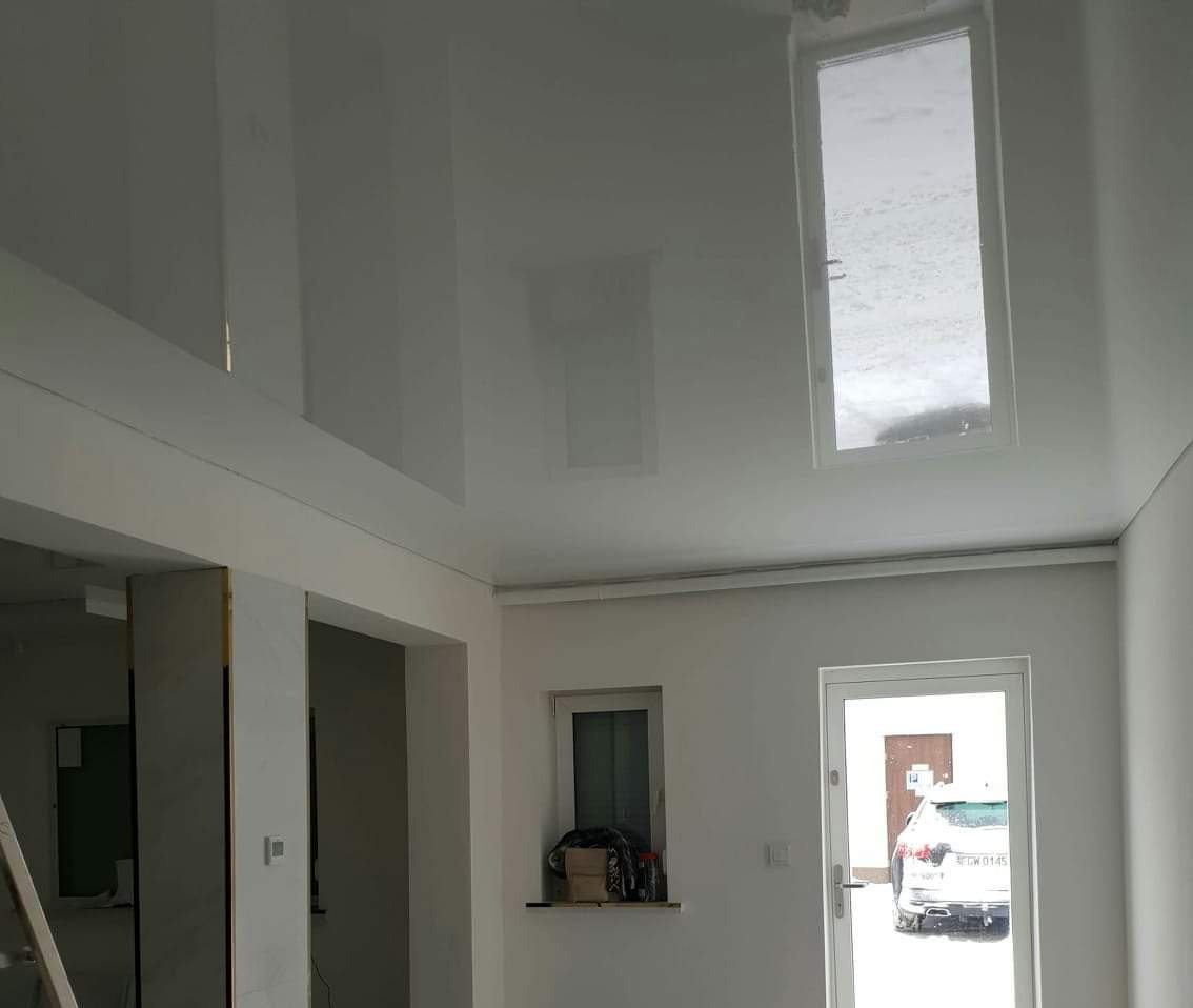 Biały, błyszczący sufit napinany z wbudowanymi oknami dachowymi w nowo wykończonym, jasnym pomieszczeniu z widocznym fragmentem drabiny i samochodem za oknem.