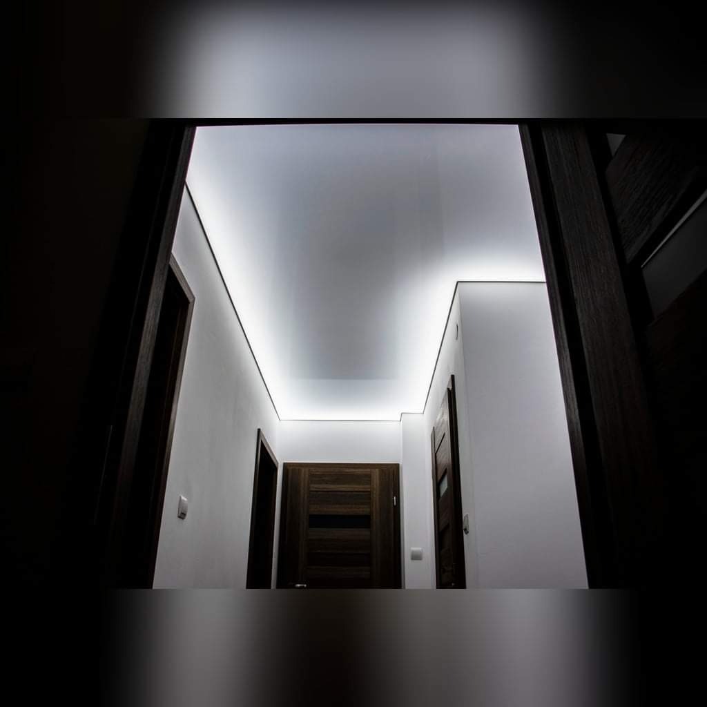 Korytarz z białym sufitem napinanym, oświetlonym listwami LED, widok z dołu, ciemne futryny drzwi po bokach.