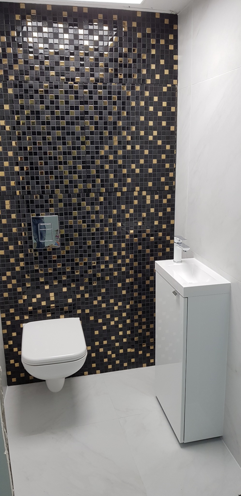 Nowoczesna łazienka z mozaikową ścianą w odcieniach czerni i złota, białą umywalką z szafką i podwieszaną toaletą.