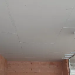 Montaż płyt kartonowo-gipsowych na suficie w surowym wnętrzu, widoczne profile mocujące, ślady po wkrętach i fragment ściany z czerwonej cegły dziurawki.