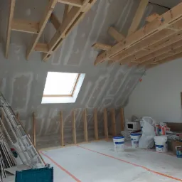 Poddasze w trakcie adaptacji: widoczne drewniane krokwie, ocieplenie wełną mineralną pod płytami gipsowo-kartonowymi, okno dachowe i narzędzia pracy na podłodze, zarys instalacji elektrycznej...