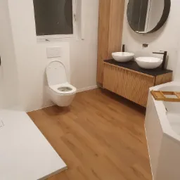 Nowoczesna łazienka z kabiną prysznicową typu walk-in, wiszącą toaletą, drewnianą szafką z umywalkami nablatowymi i okrągłym lustrem.