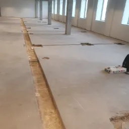 Betonowa posadzka w trakcie remontu biura, z wyciętym rowem na instalacje i workiem zaprawy obok, widoczne słupy i okna.