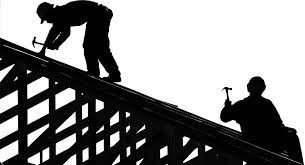 Sylwetki dwóch dekarzy pracujących na dachu, jeden wbija gwóźdź młotkiem, drugi trzyma młotek w dłoni, widoczne elementy konstrukcji dachu na tle jasnego nieba.