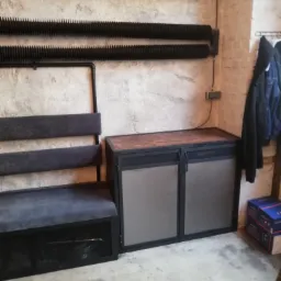 Loftowa aranżacja wnętrza z tapicerowaną ławką z oparciem i szafką z drewnianym blatem na metalowym stelażu, na tle surowej ściany z grzejnikami.