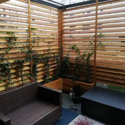 Zadaszony taras z drewnianą konstrukcją żaluzjową, po której pną się rośliny, oświetlony girlandą świetlną, z meblami wypoczynkowymi i stolikiem kawowym.