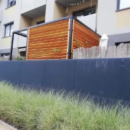 Drewniana konstrukcja z poziomych listew, tworząca osłonę tarasu lub balkonu, z pnączami roślin, widziana z poziomu trawy przed budynkiem mieszkalnym.