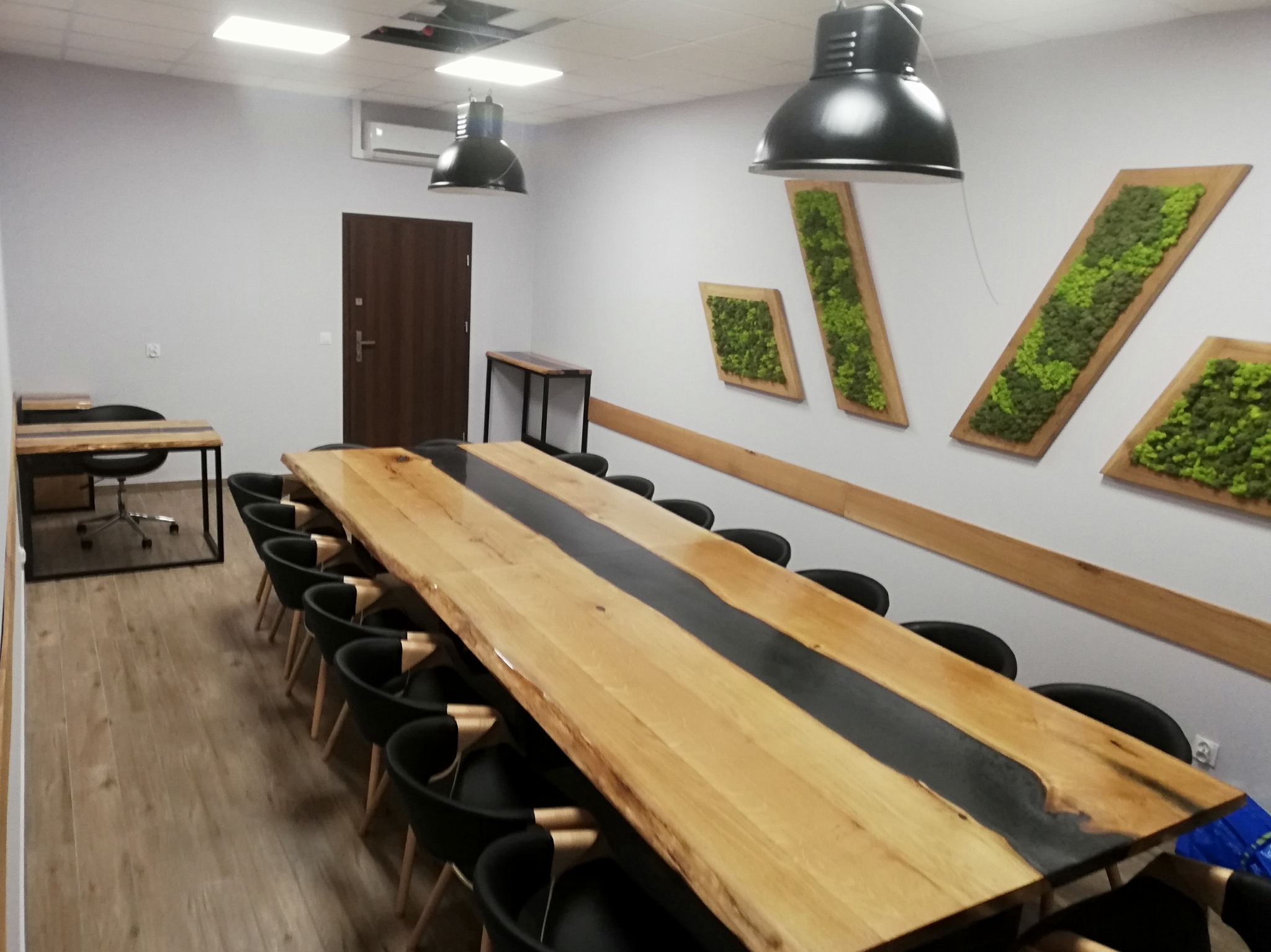 Długi, drewniany stół z żywicą epoksydową w nowoczesnej sali konferencyjnej, otoczony czarnymi krzesłami, z dekoracjami z mchu na ścianie i lampami wiszącymi.