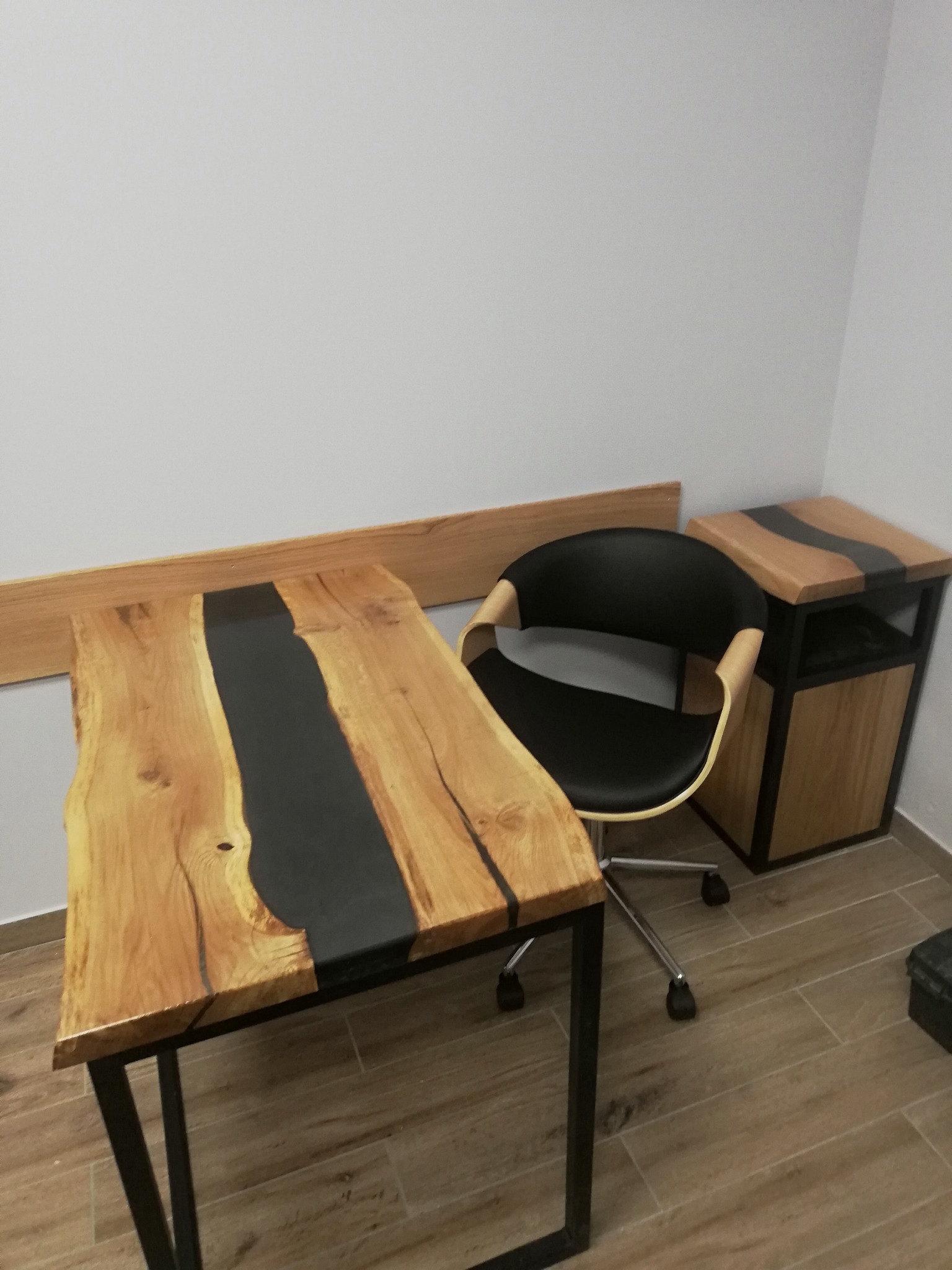 Biurko z blatem z drewna i żywicy epoksydowej w kolorze czarnym, obok krzesło biurowe i szafka nocna z podobnym blatem, całość w minimalistycznym wnętrzu.