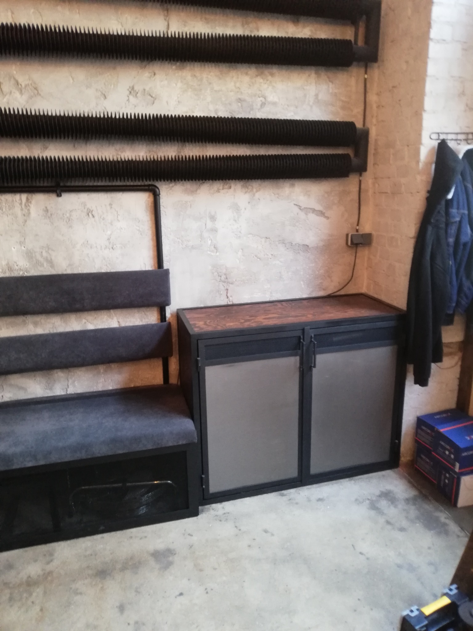 Loftowa aranżacja wnętrza z tapicerowaną ławką z oparciem i szafką z drewnianym blatem na metalowym stelażu, na tle surowej ściany z grzejnikami.