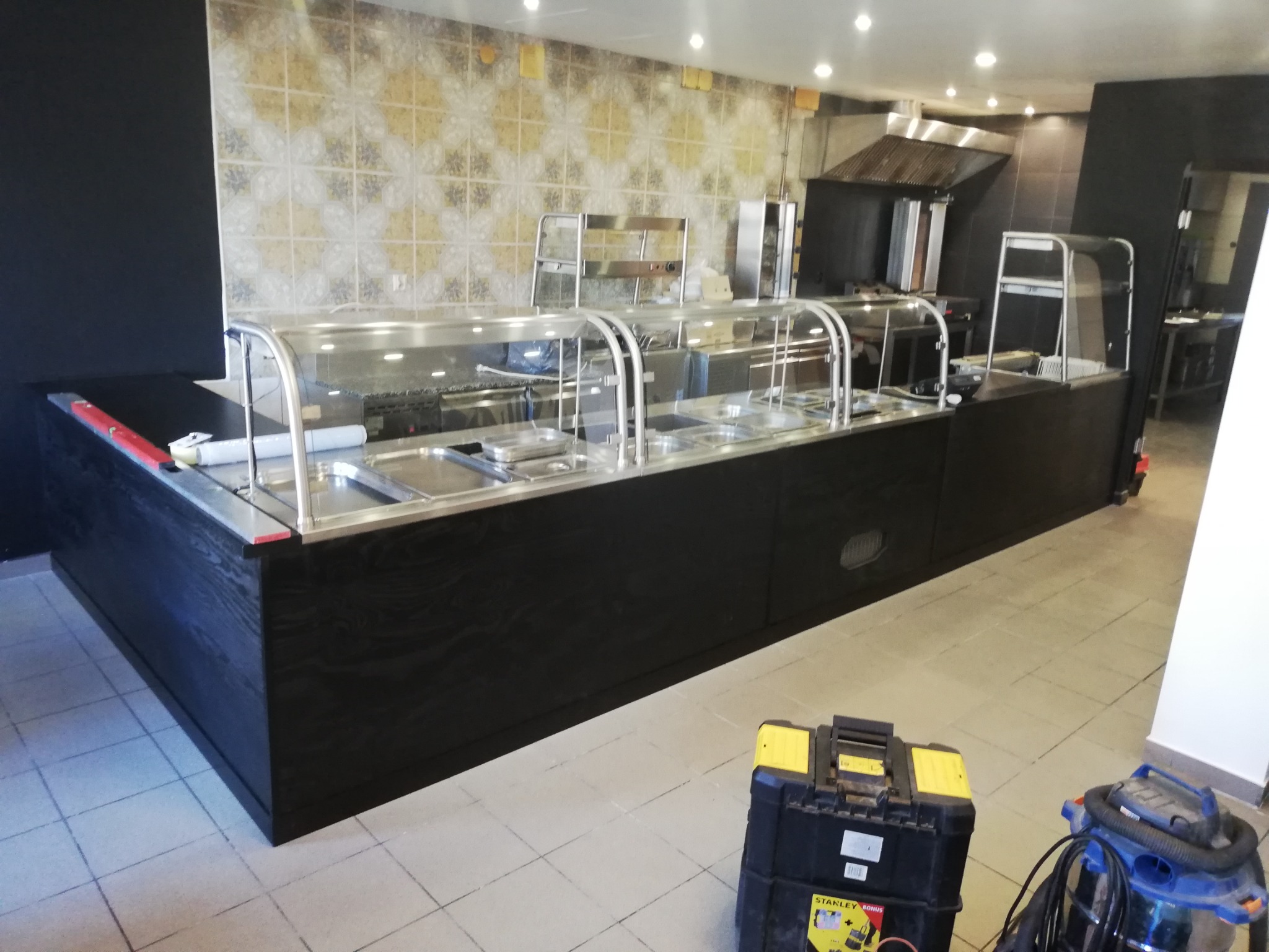 Częściowo zainstalowana lada chłodnicza ze stali nierdzewnej i czarnego drewna w lokalu gastronomicznym, widoczna skrzynka narzędziowa i odkurzacz przemysłowy na podłodze.