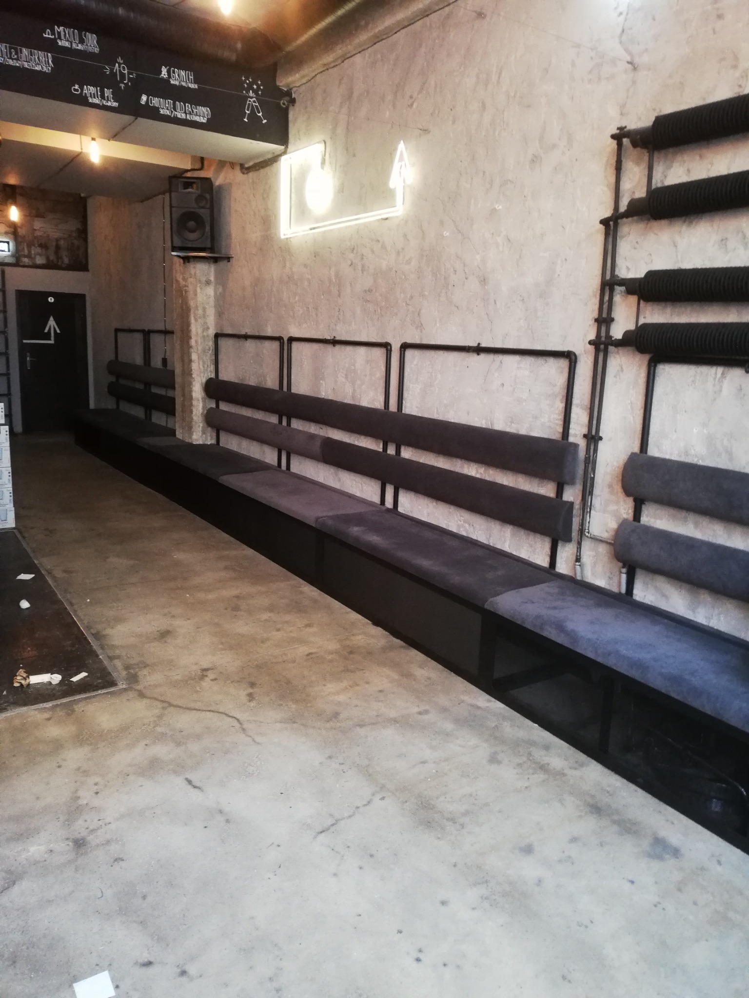 Wnętrze lokalu z długą, tapicerowaną ławką pod ścianą z szarego betonu, nad którą wisi neon w kształcie strzałki i tablica z menu drinków.