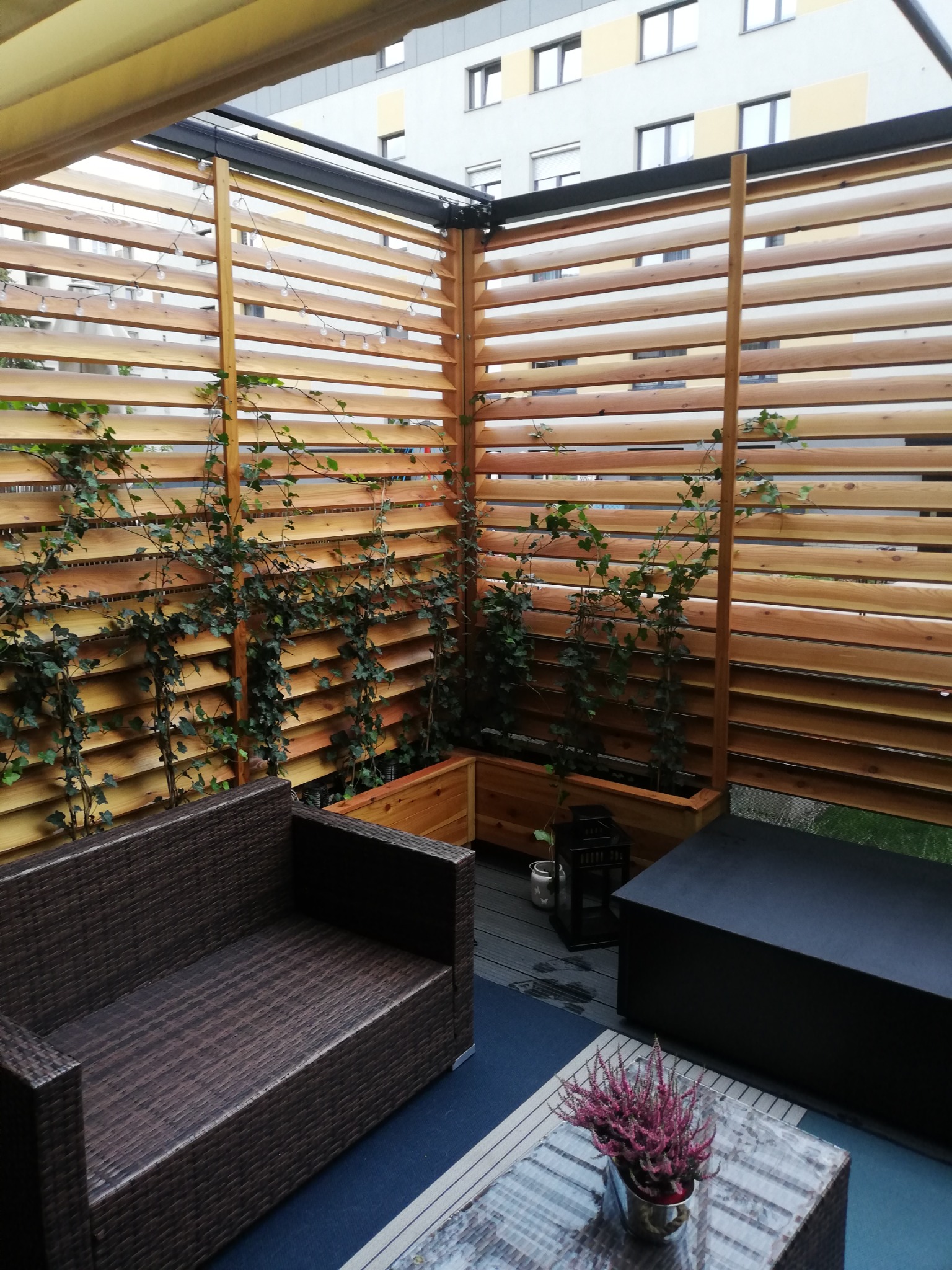 Zadaszony taras z drewnianą konstrukcją żaluzjową, po której pną się rośliny, oświetlony girlandą świetlną, z meblami wypoczynkowymi i stolikiem kawowym.