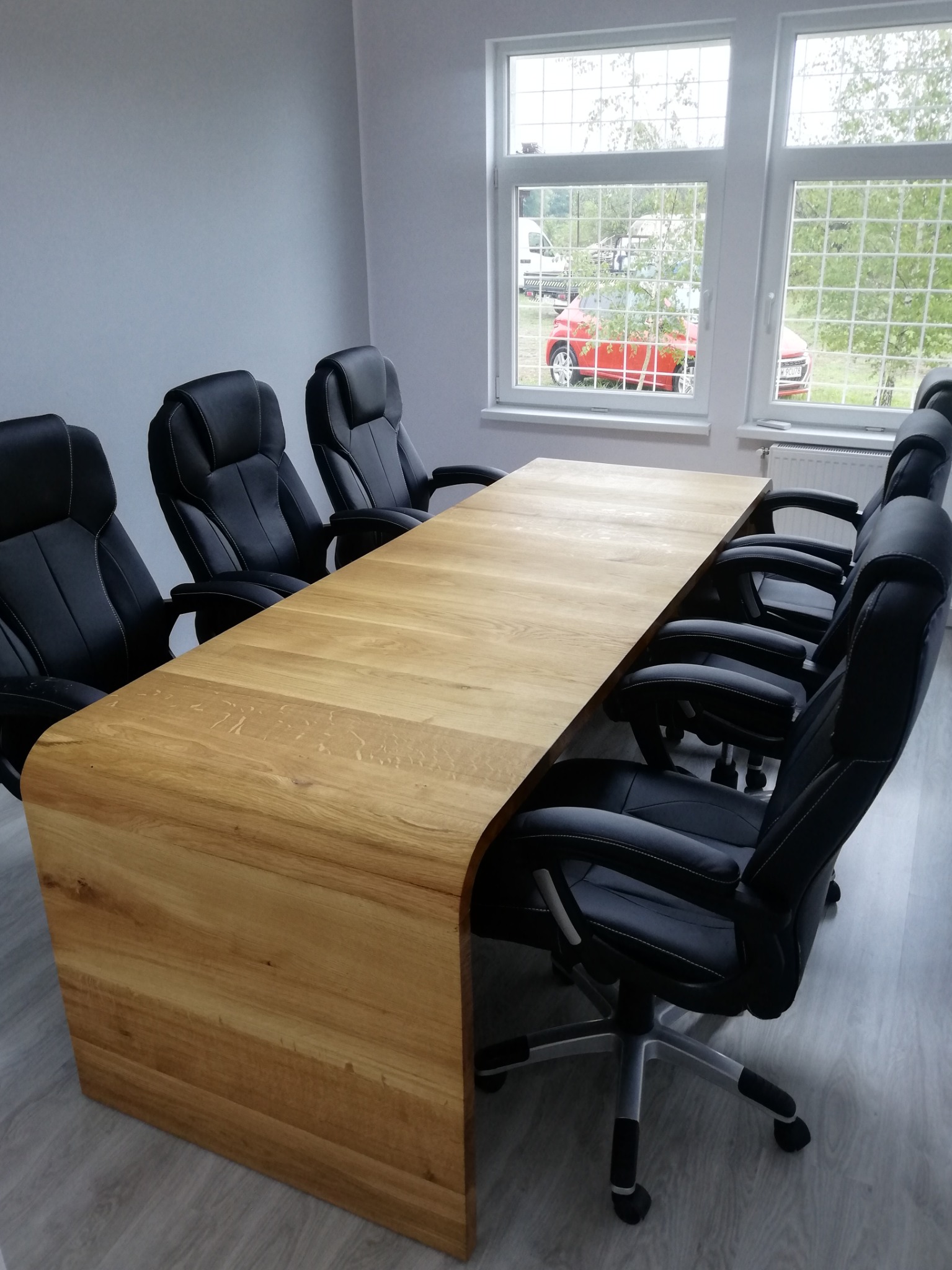 Stół konferencyjny z blatem z jasnego drewna, otoczony czarnymi krzesłami biurowymi na kółkach, widok z boku, w tle okno z widokiem na zieleń, Wrocław.
