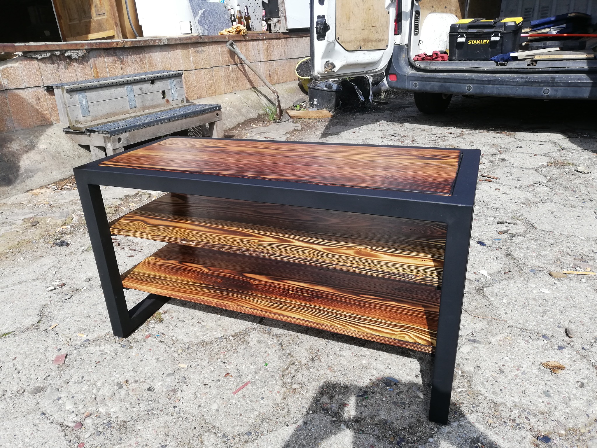 Ręcznie robiona półka z drewna o wyraźnym usłojeniu i czarnej metalowej ramie, sfotografowana na zewnątrz w surowym otoczeniu z narzędziami i samochodem dostawczym w tle.