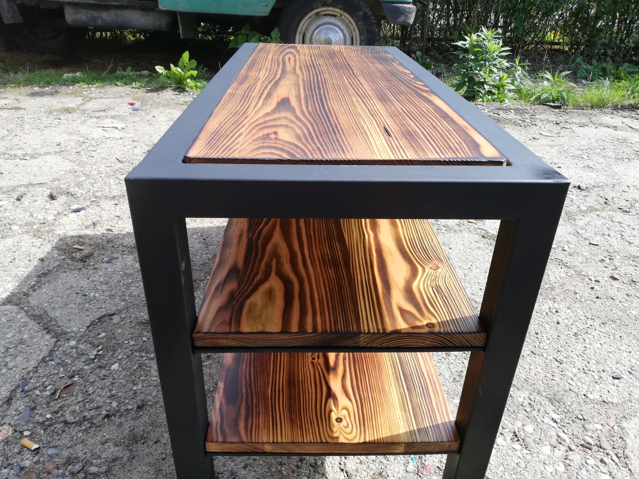 Ręcznie robiony regał z trzema półkami z opalizowanego drewna na czarnej metalowej ramie, ustawiony na żwirowym podłożu z fragmentem zielonego pojazdu w tle.