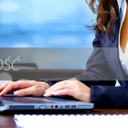 Kobieta w marynarce pracuje na laptopie przy biurku z notatnikiem, widoczne szklanki z wodą, w tle rozmyte biuro.