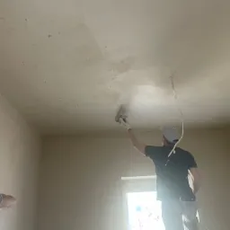 Pracownik w trakcie nakładania tynku na sufit w pomieszczeniu, widoczne niedociągnięcia i nierówności na powierzchni, kabel zwisający z sufitu.
