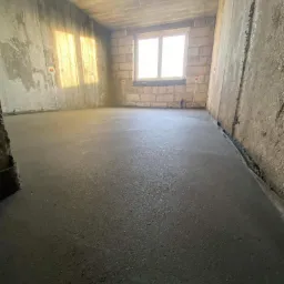 Świeżo wylana, gładka posadzka w surowym pomieszczeniu z oknem, widoczne ślady po instalacjach elektrycznych na ścianach.