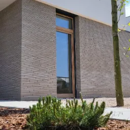 Nowoczesna elewacja budynku z szarej cegły klinkierowej ułożonej poziomo, widoczne okno z drewnianą ramą, fragment drzewa i rabata wysypana korą.