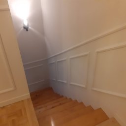 Sławomir Jurga - Klatka schodowa z drewnianymi schodami i ścianami z białymi panelami dekoracyjnymi oraz oświetleniem punktowym.