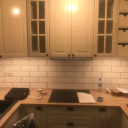 Sławomir Jurga - Jasna kuchnia z białymi płytkami metra, drewnianym blatem i czarnym zlewem, przedłużona lampa sufitowa, butelka wody i telefon na blacie.