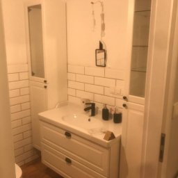 Sławomir Jurga - Biała łazienka z umywalką nablatową, szafką z szufladami, wysoką szafką z przeszklonym frontem i białymi płytkami na ścianie, oświetlenie w trakcie montażu.