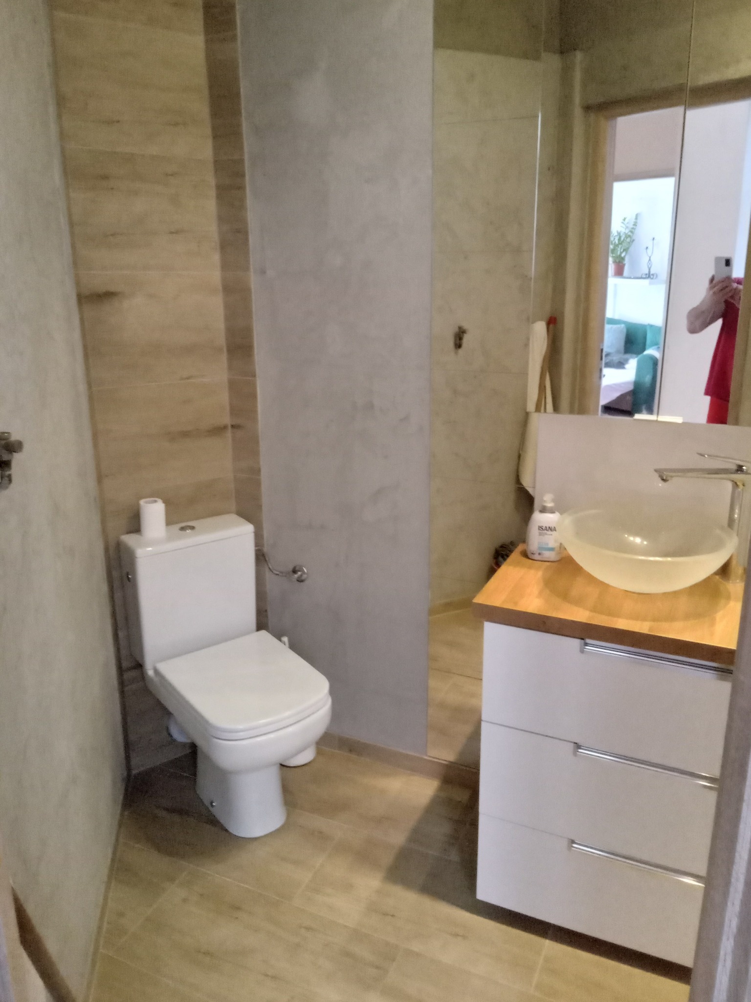 Nowoczesna łazienka z toaletą, umywalką na szafce z drewnianym blatem i lustrem, w którym odbija się osoba robiąca zdjęcie.