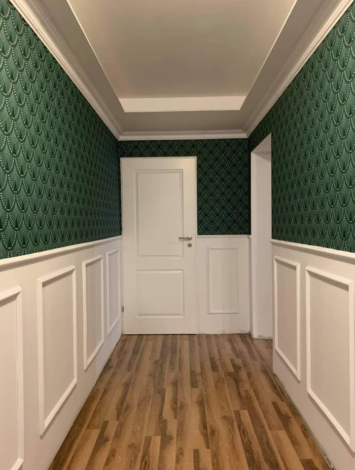 Korytarz z zieloną tapetą w stylu art déco, białą boazerią i drewnopodobną podłogą, oświetlony górnym światłem z sufitu.