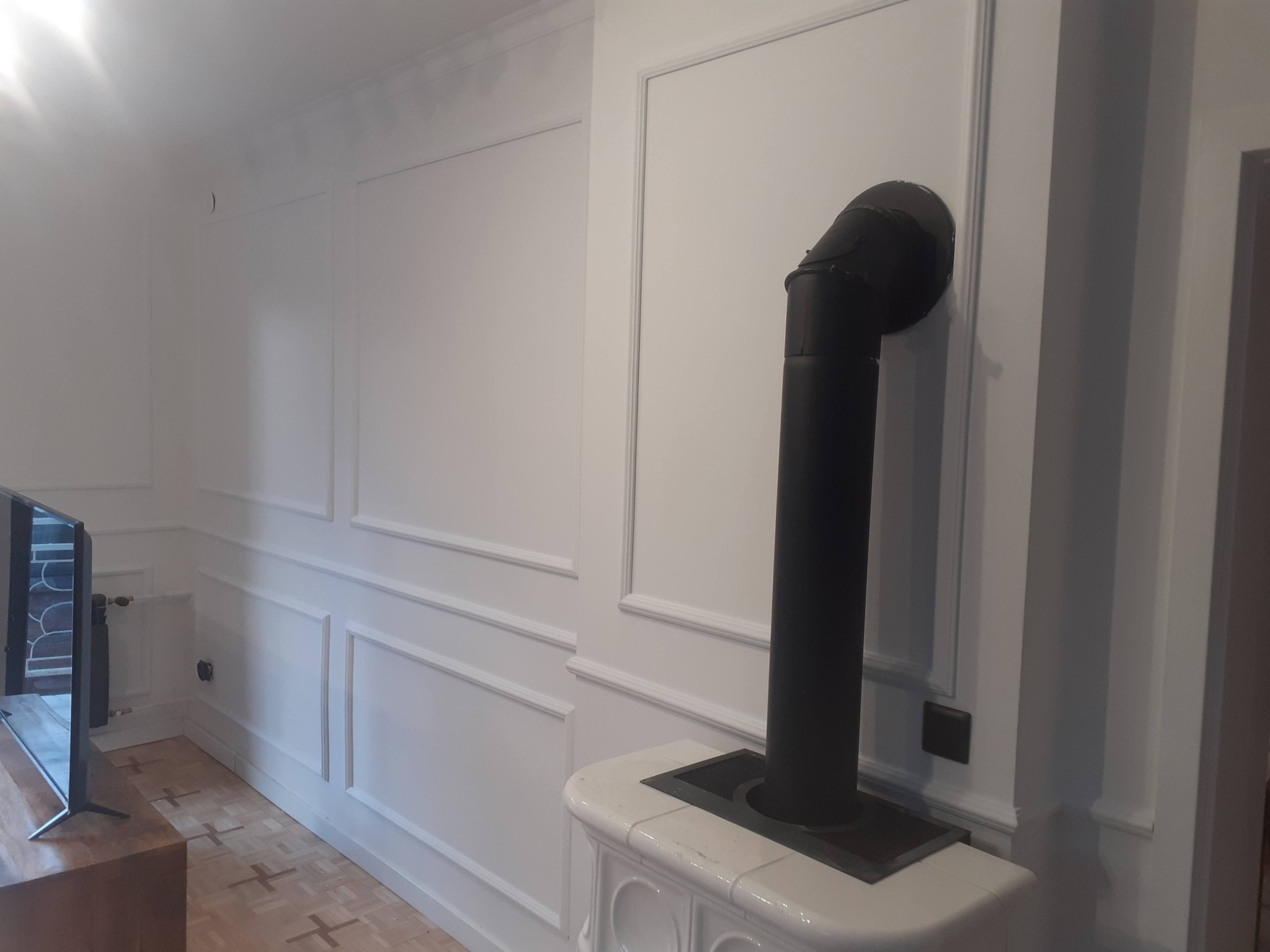 Białe ściany z dekoracyjnymi listwami, piec kaflowy z czarną rurą odprowadzającą dym, parkiet w geometryczny wzór, widoczny telewizor.