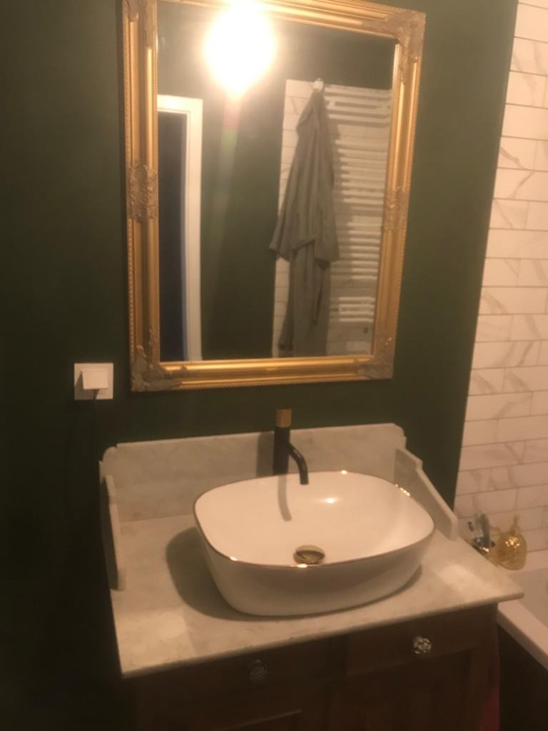 Elegancka umywalka nablatowa z czarną armaturą na marmurowym blacie, pod ozdobnym lustrem w złotej ramie na tle ciemnozielonej ściany i białych płytek.