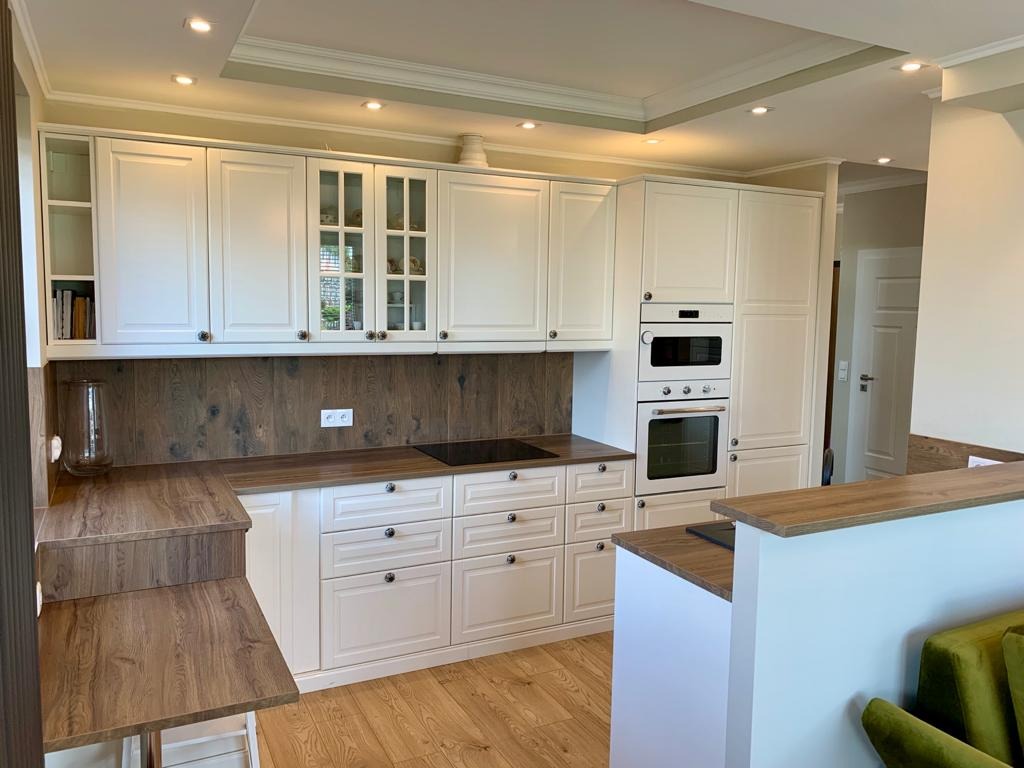 Jasna kuchnia w stylu klasycznym z białymi szafkami, drewnianym blatem i panelami ściennymi, widok na salon z zieloną sofą.